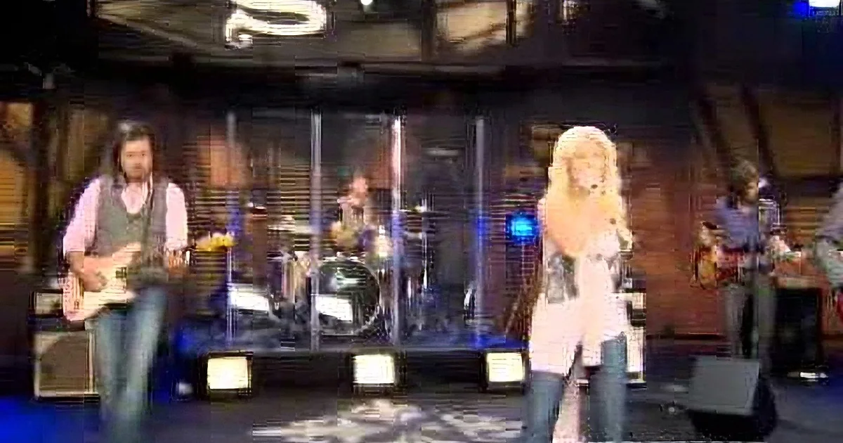 A musician plays guitar on stage, surrounded by band members and a singer, with a vibrant stage setup and lighting.