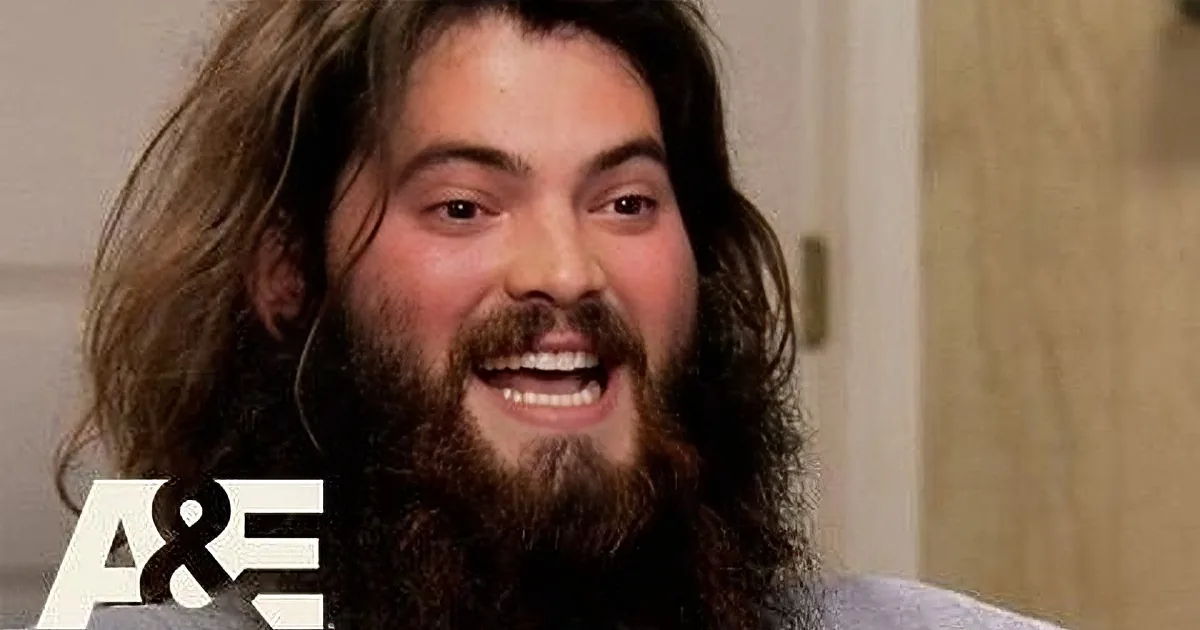 A man with long hair and a beard smiles widely, wearing a light-colored shirt, with a beige wall and A&E logo in the...