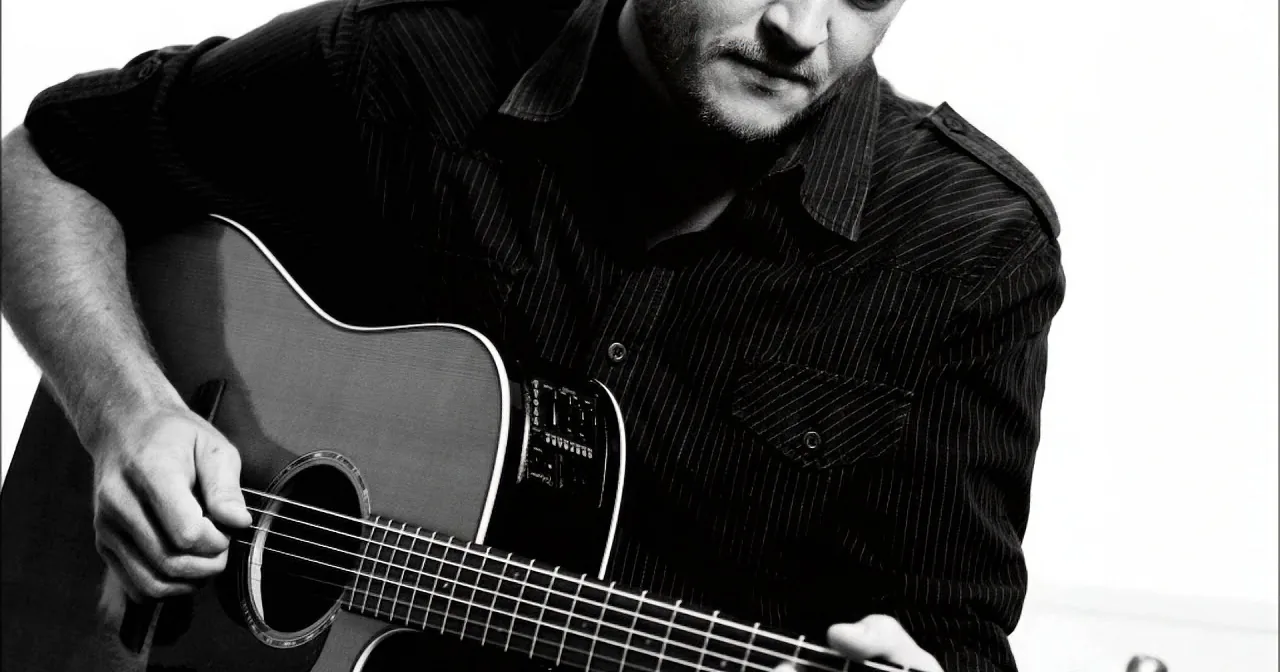 A man in a striped shirt plays an acoustic guitar, focusing on his fingers on the strings.