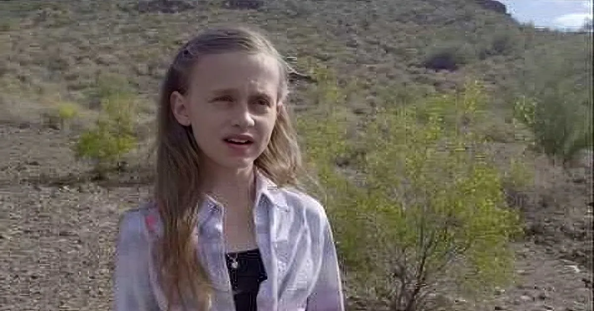 A young girl with long blonde hair stands outdoors in a desert-like environment, wearing a white shirt over a black top.