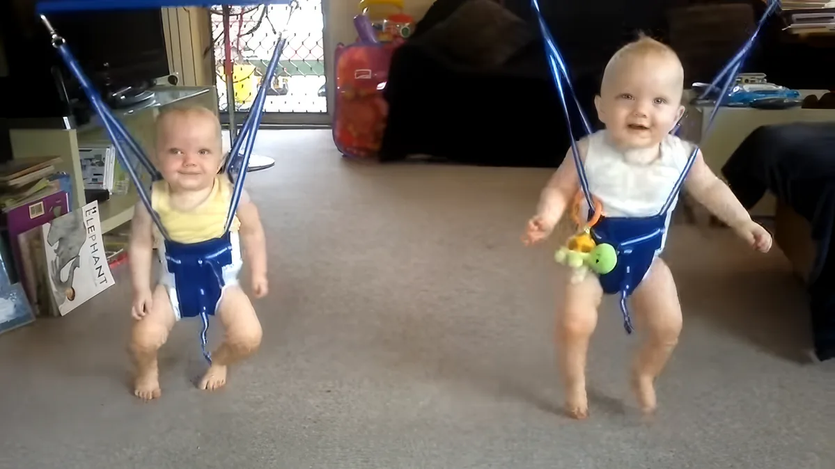 A baby is swinging in a blue baby swing, wearing a white top and blue pants, with a toy attached to the swing.