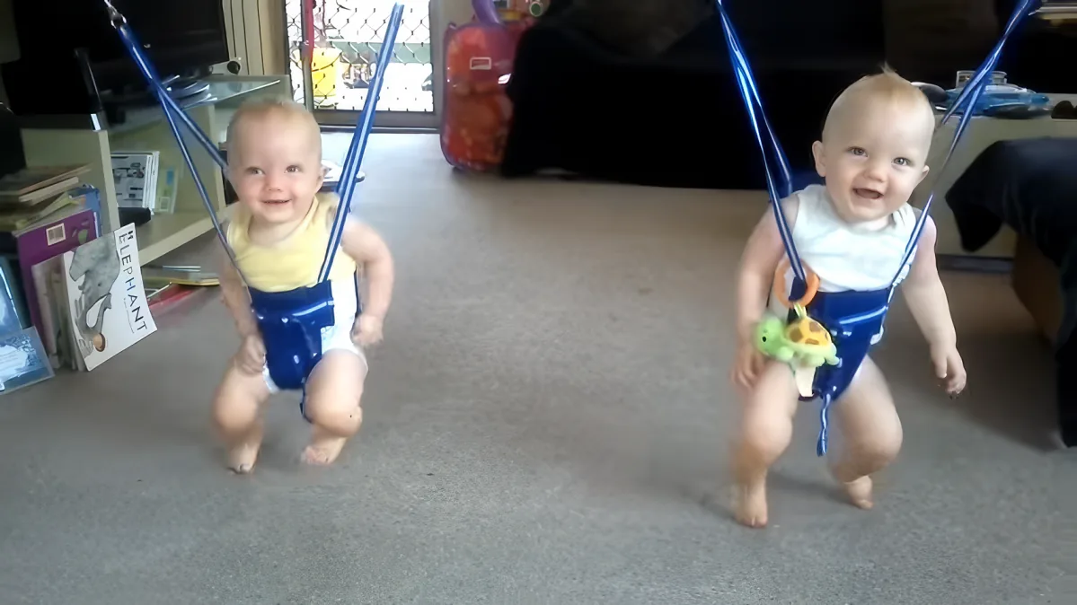 Two babies in baby bouncers, one in yellow, one in white, both smiling and bouncing.