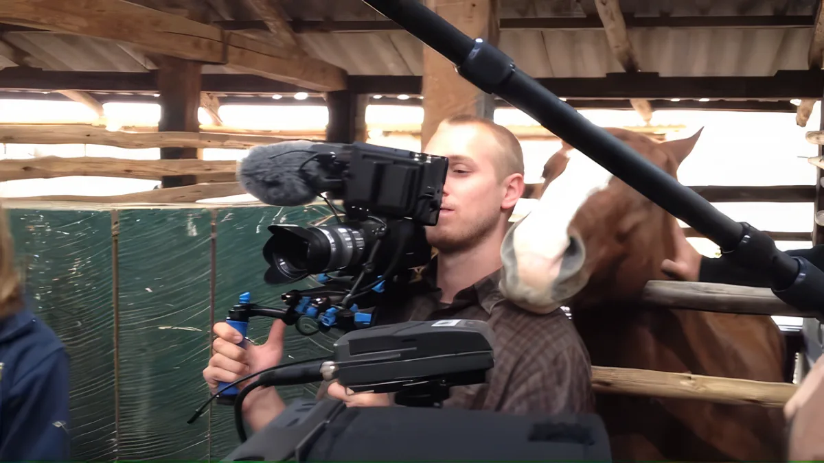 A man films a horse in a barn using a large camera with a microphone attached.