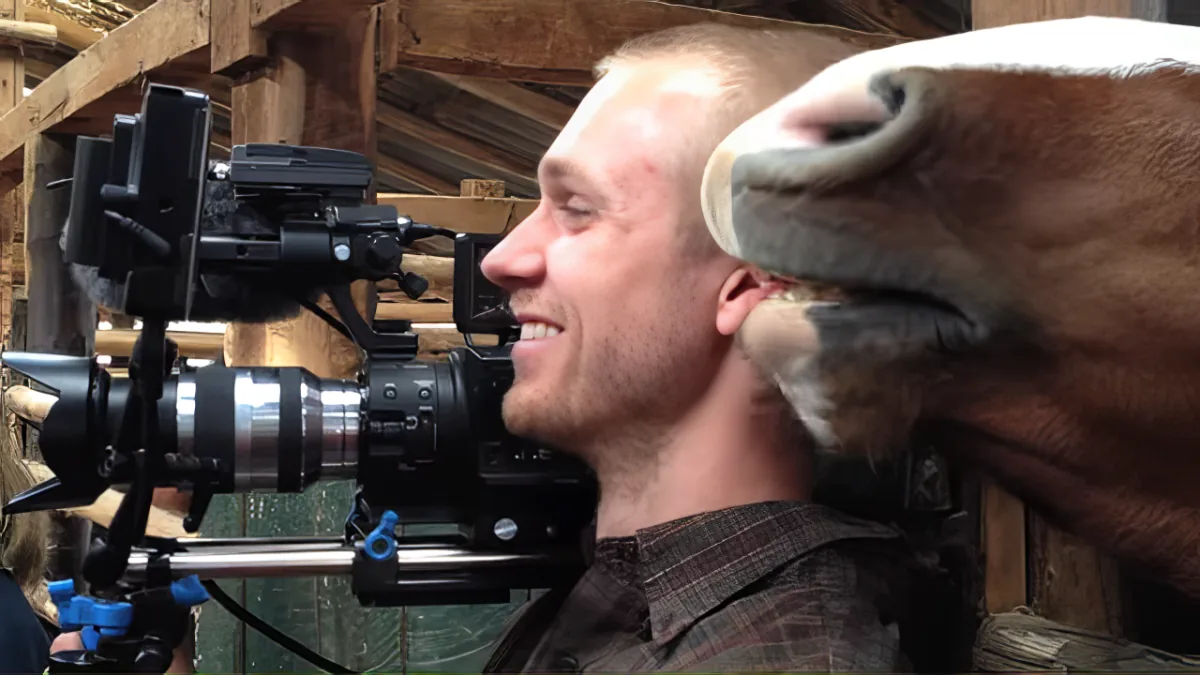 A man smiles while filming a horse with a camera rig in a barn setting.