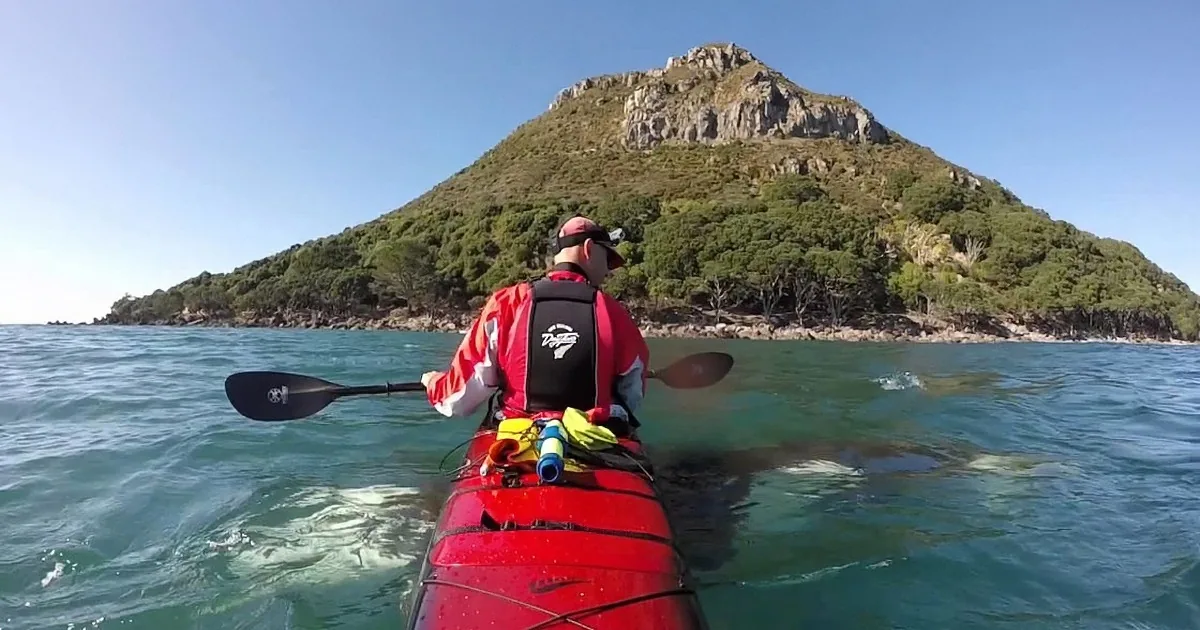 A person paddles a red kayak on calm water, with a rocky island and lush greenery in the background.