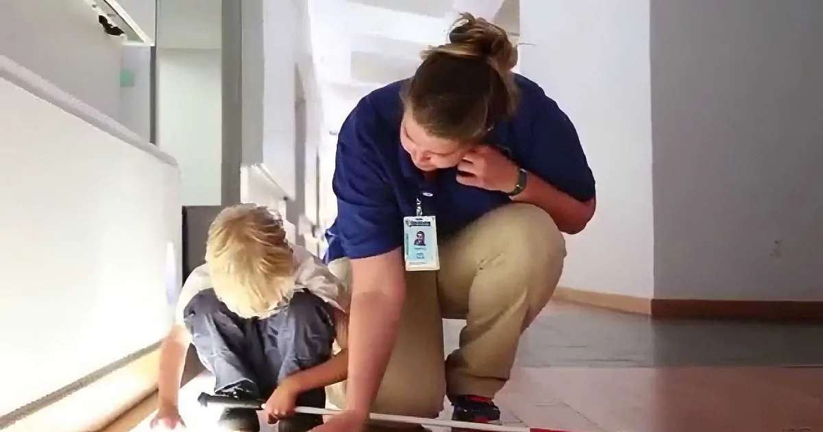 A woman in a blue shirt and khaki pants kneels on a wooden floor, interacting with a child who is also kneeling nearby.