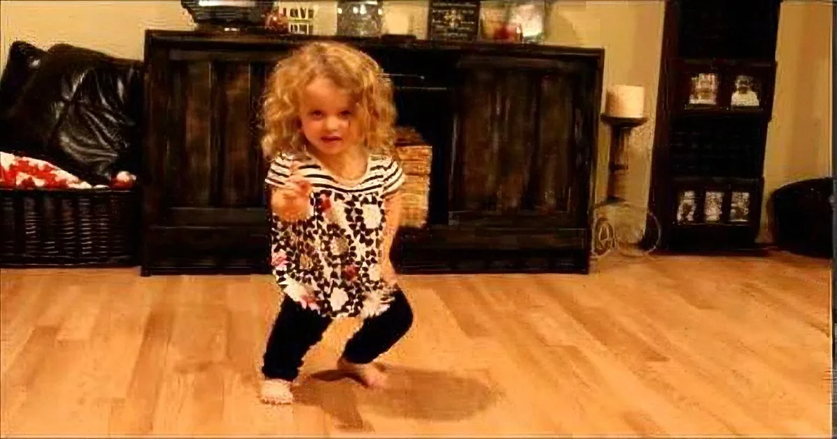 A toddler in a striped shirt and black pants stands on a wooden floor, holding something in her hand.