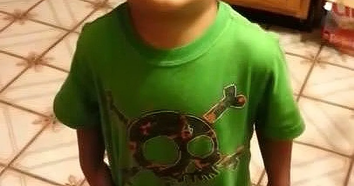 A child in a green t-shirt with a skull design stands in a kitchen with tiled flooring.