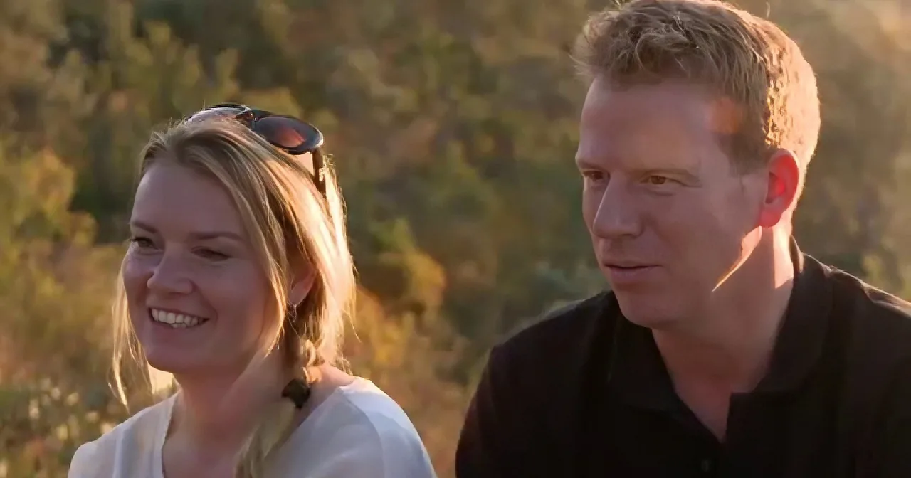 A man in a black shirt and a woman in a white top are smiling outdoors, with a blurred background of greenery.
