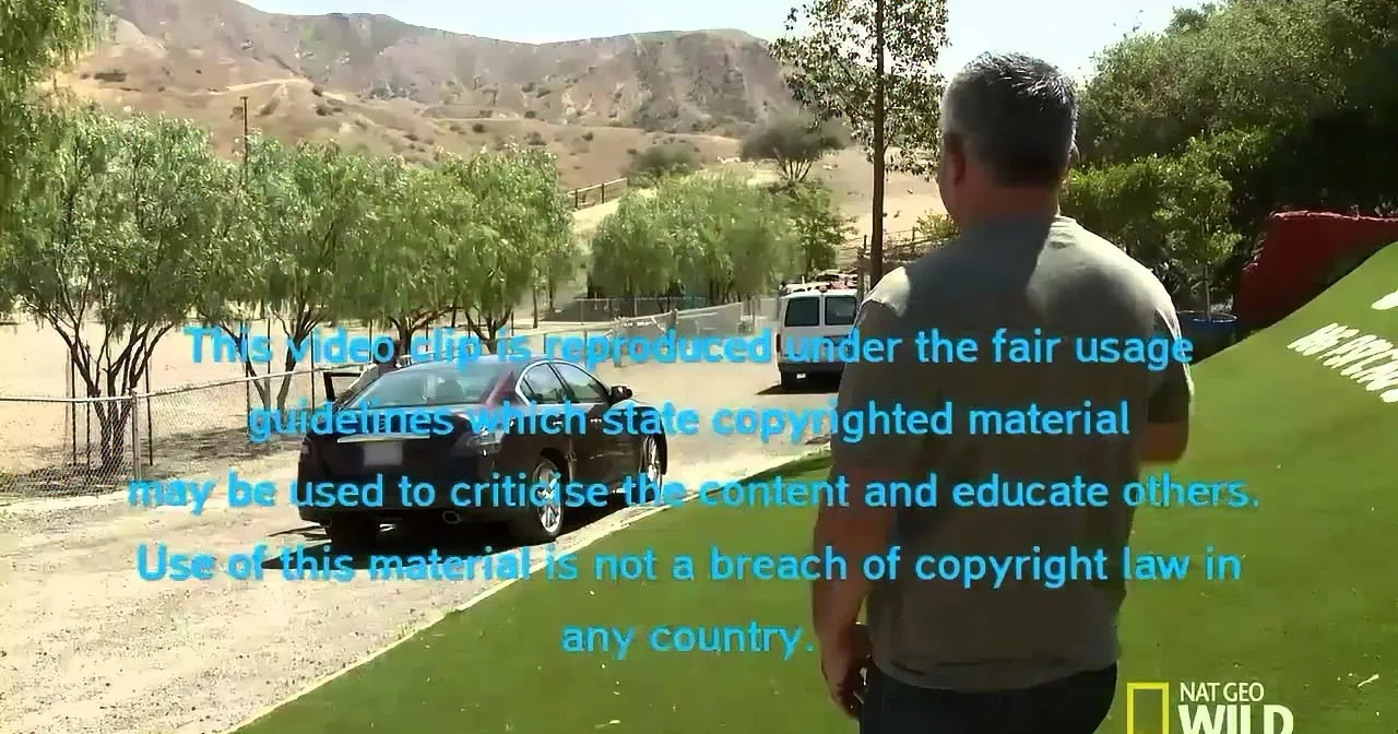 A man walks on a grassy path near a parking lot with a car and a van, surrounded by trees and hills.