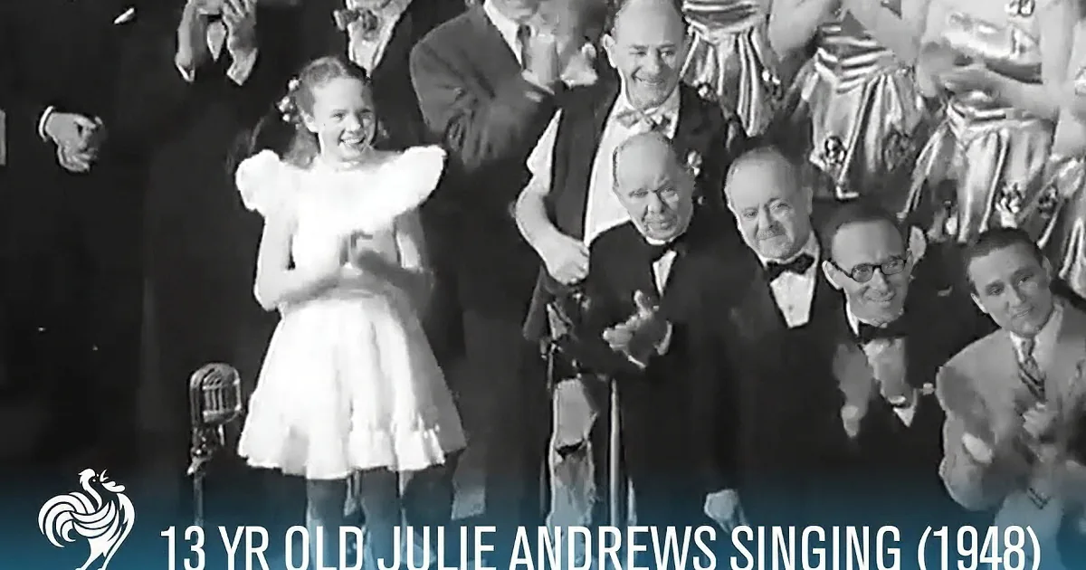 A young girl in a white dress is smiling on stage, surrounded by adults in formal attire, with text indicating she's...