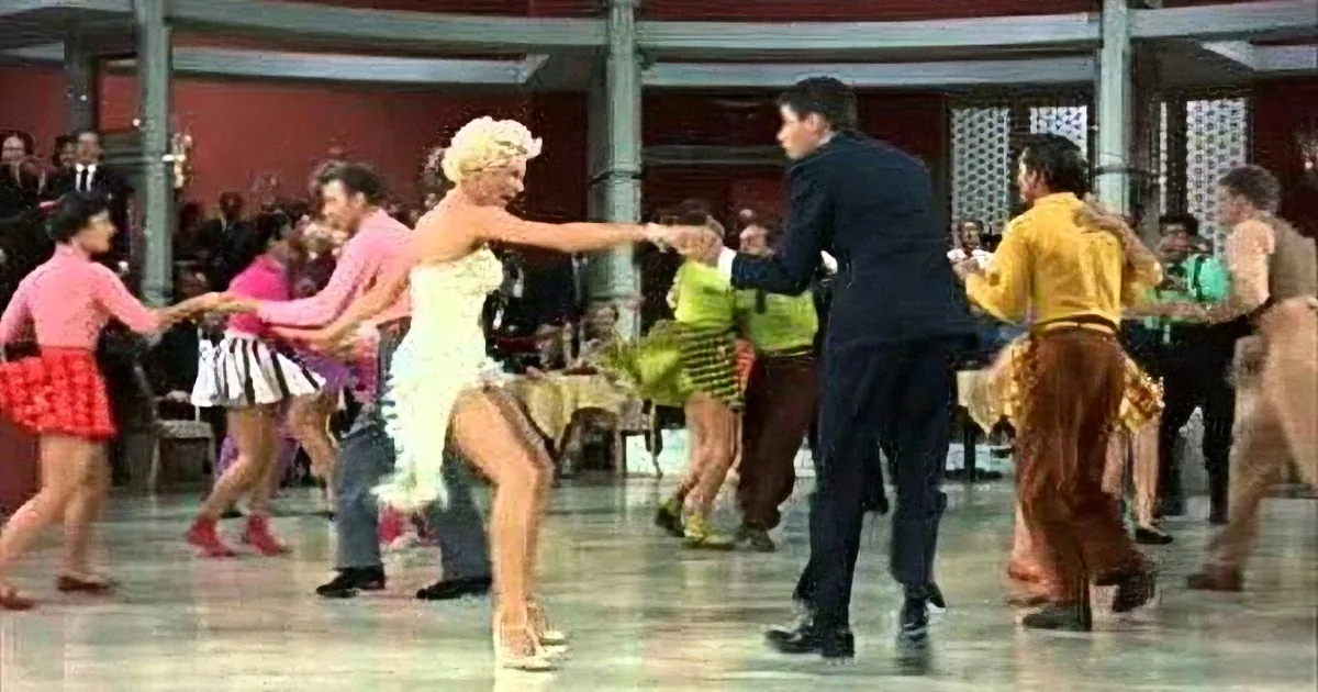 A woman in a white dress dances energetically with a man in a dark suit in a lively ballroom setting.