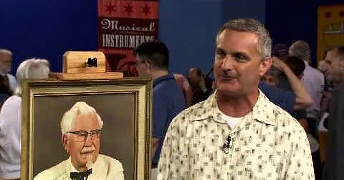 A man in a patterned shirt stands next to a portrait of Colonel Sanders, with a crowd and musical instruments in the...