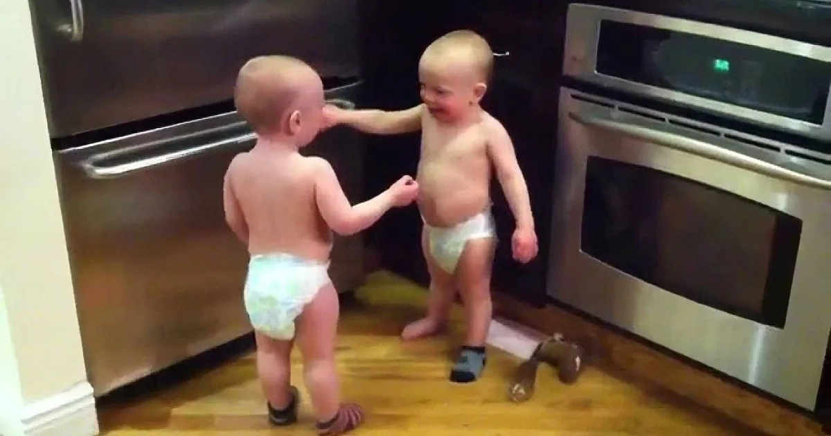 Two toddlers, one in a diaper, interact near a stainless steel oven in a kitchen.