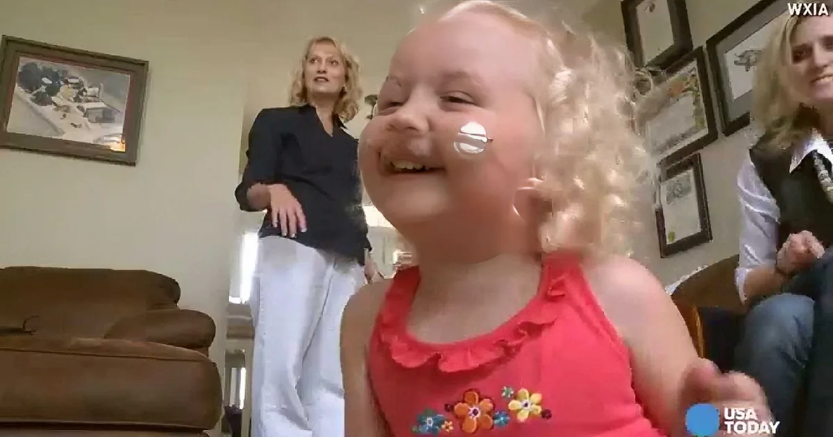 A young girl with curly blonde hair and a pink dress is smiling, with a white substance on her cheek.