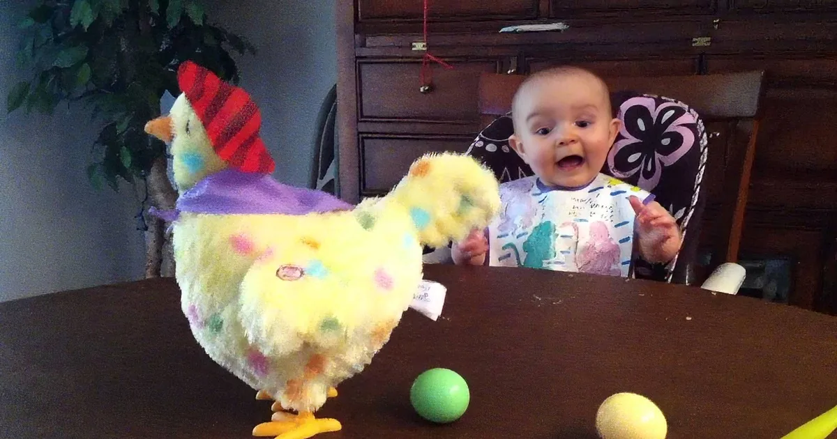 A baby in a high chair smiles as a colorful plush chicken dances on a table with Easter eggs.