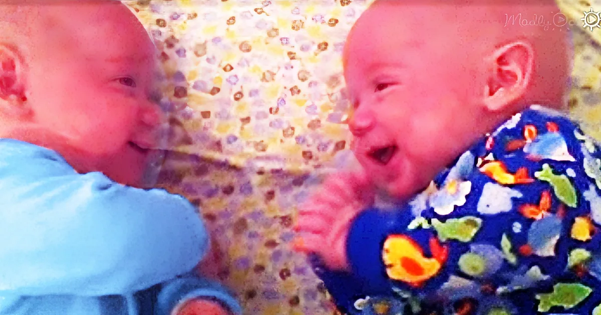 Two babies, one in blue, the other in colorful pajamas, are interacting closely, with one appearing to smile or laugh.