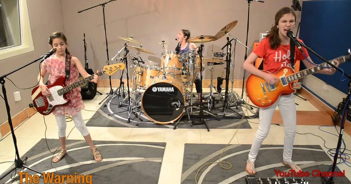 A young girl in a red shirt plays guitar while another girl in a pink dress plays bass in a music studio.