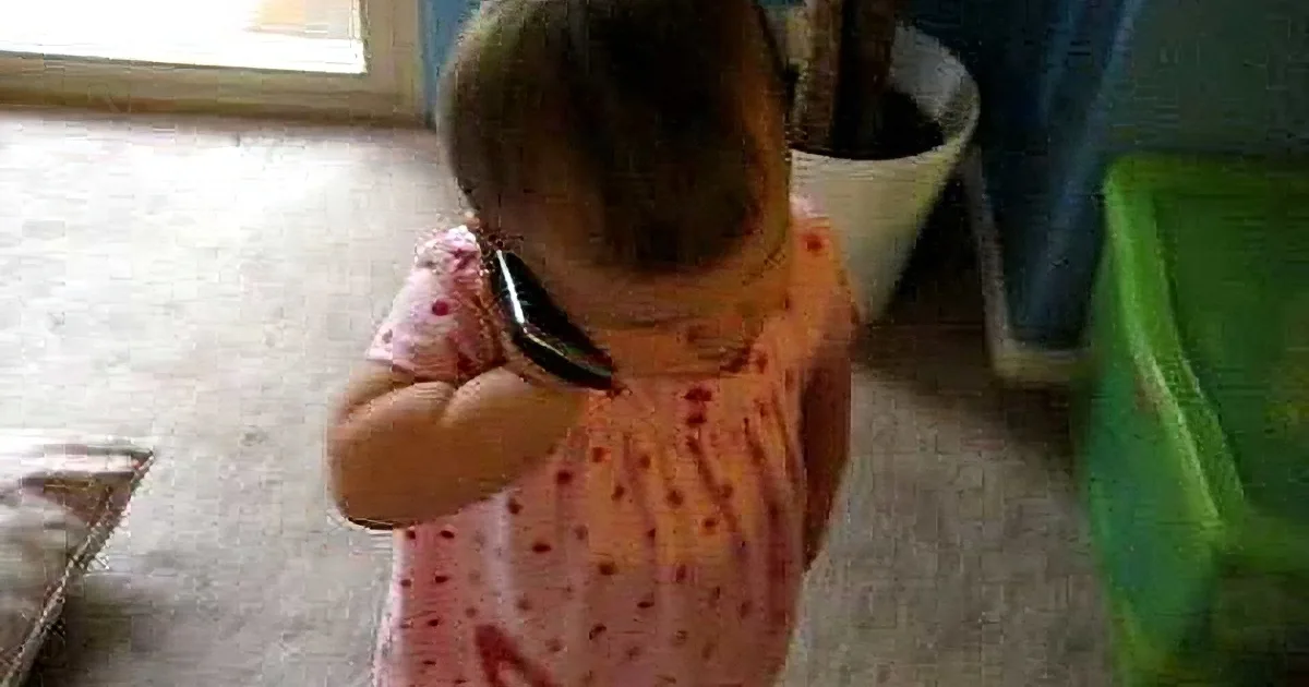 A baby in a pink dress holds a black object near their ear, possibly a phone, in a room with a potted plant and green...
