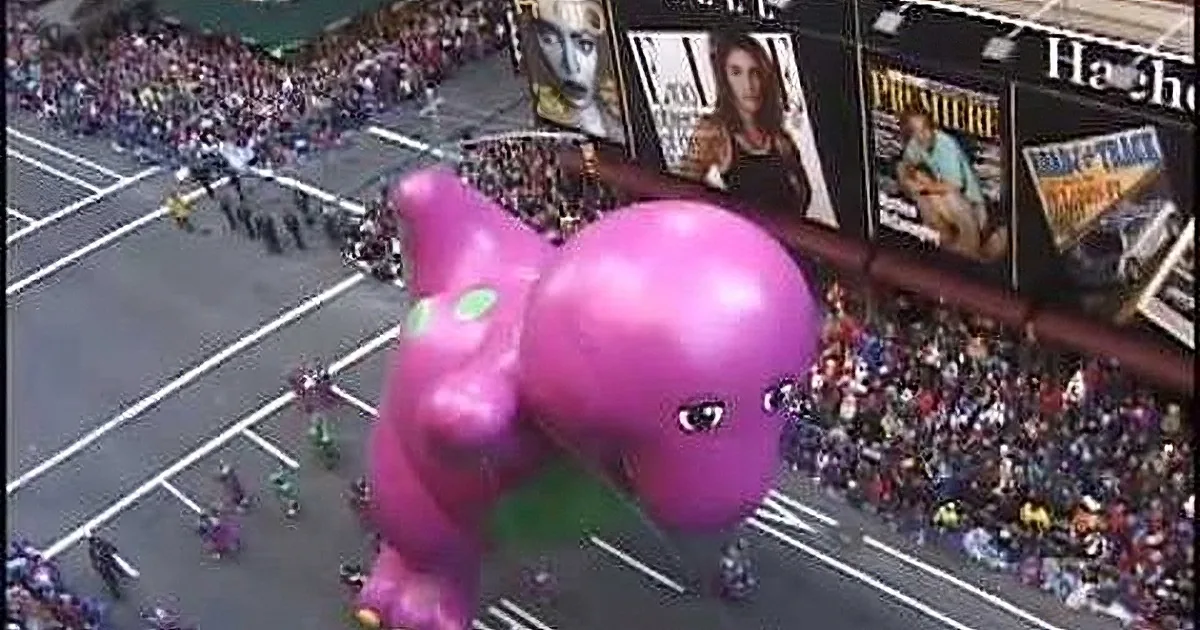 A large pink dinosaur balloon floats above a busy street during a parade, with spectators lining the sides.