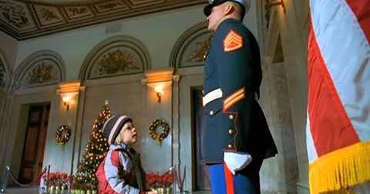 A young child in a red jacket stands in front of a Christmas tree, looking up at a uniformed officer in a grand room.