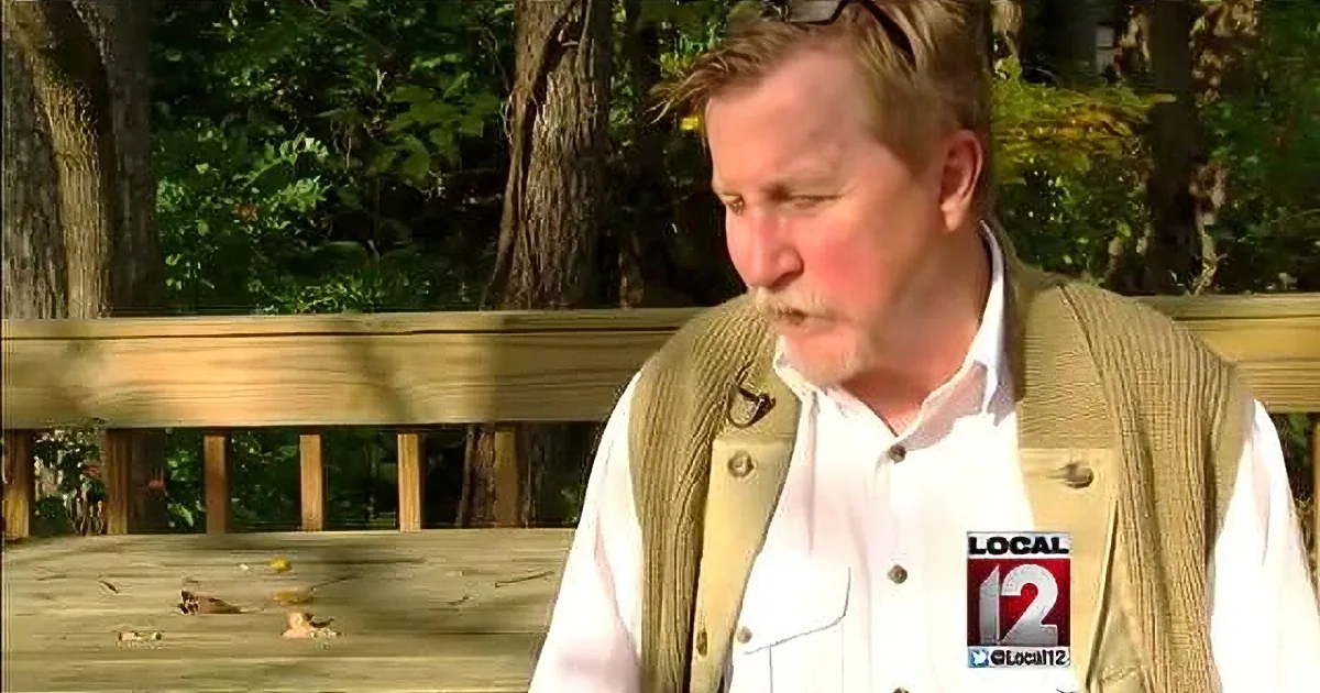 A man in a white shirt and beige vest sits on a wooden bench outdoors, surrounded by trees.
