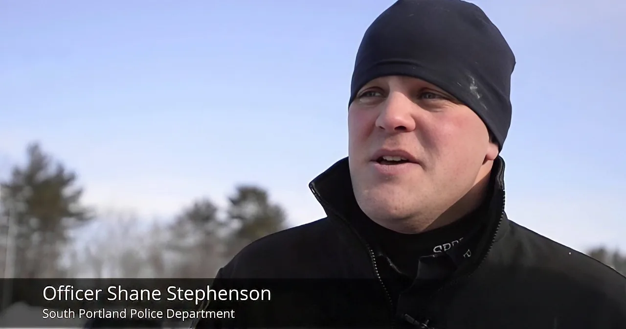 Officer Shane Stephenson from the South Portland Police Department is speaking outdoors, wearing a black jacket and beanie.