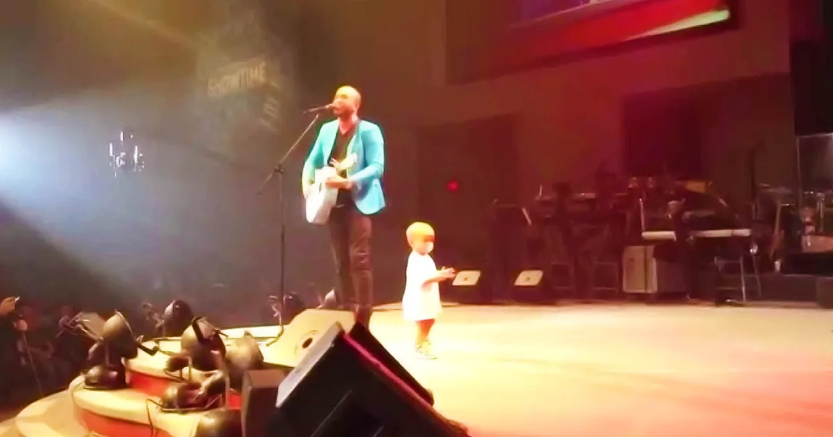 A man in a blue jacket plays guitar on stage, accompanied by a young child in a white dress.