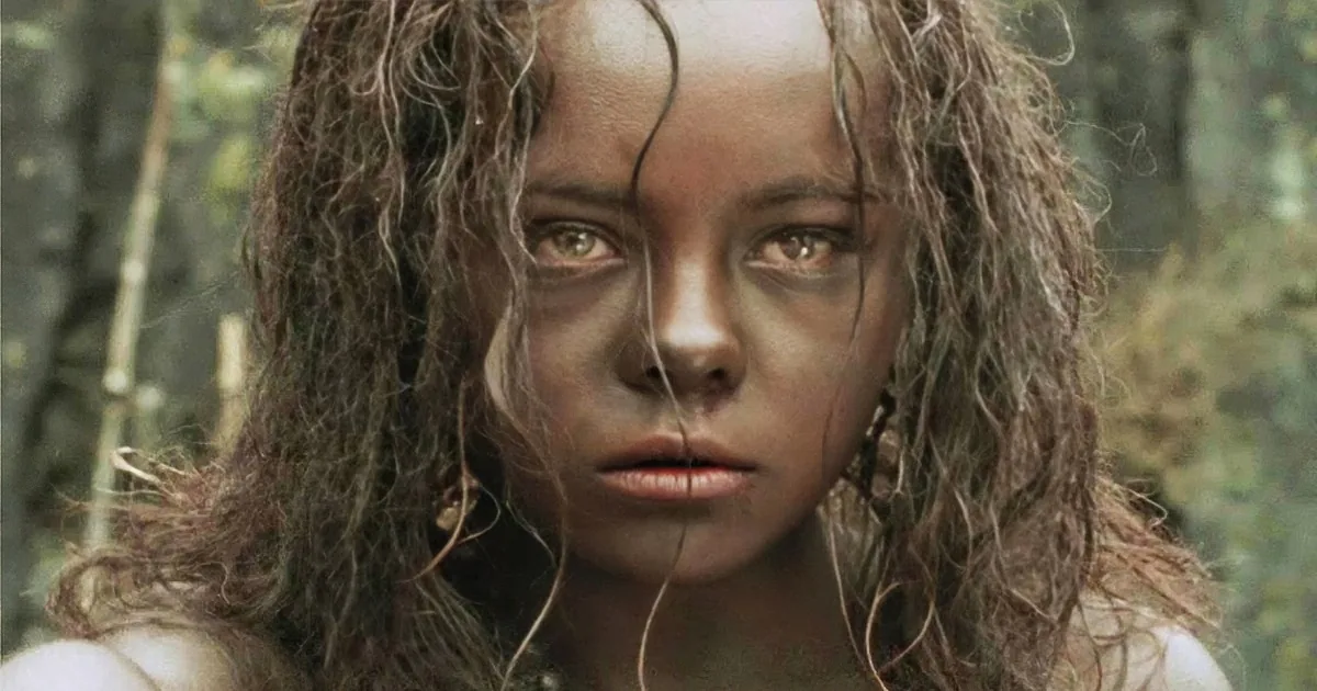 A person with wet, curly hair and a pale complexion, standing in a forested area.