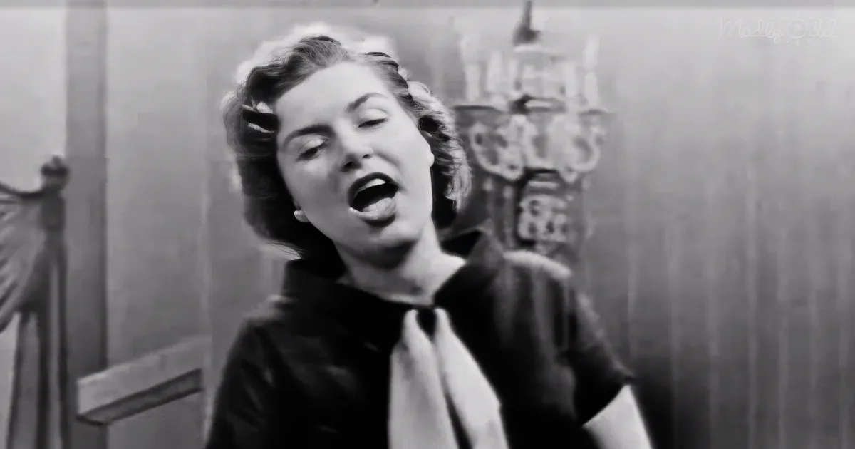 A woman in a black outfit with a white scarf is singing passionately in a room with a chandelier.