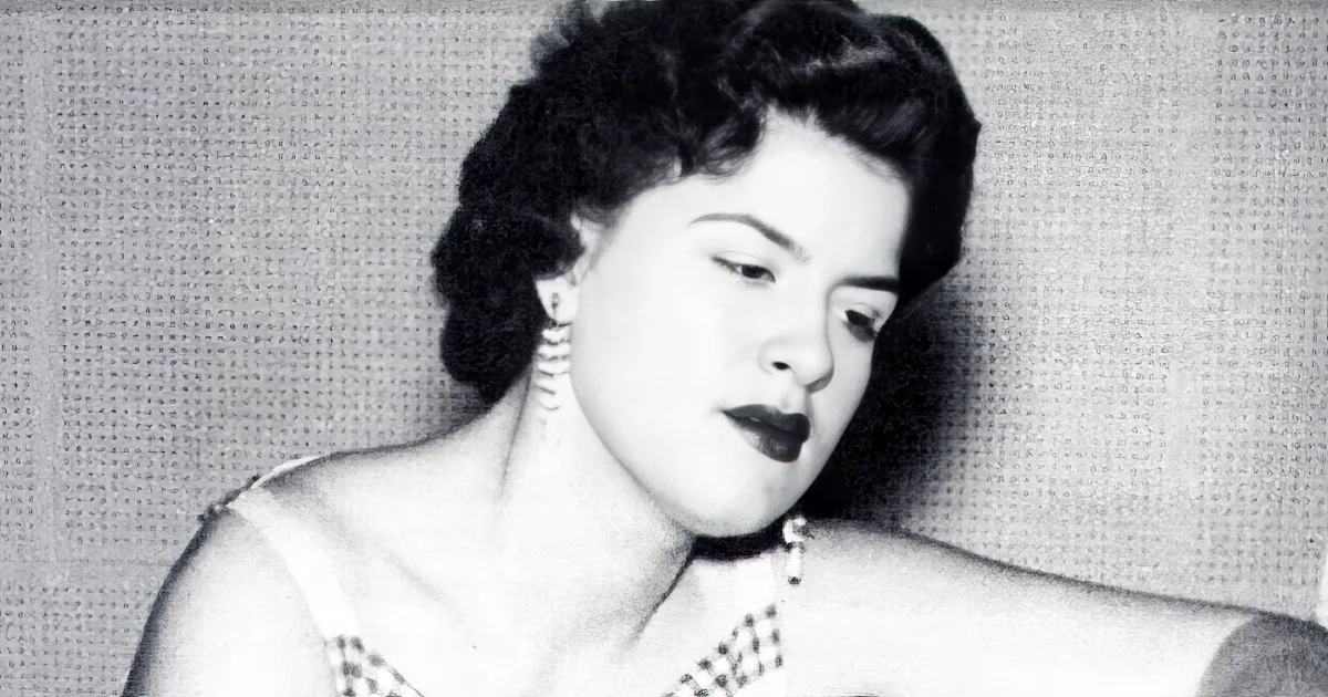 A woman with dark hair, wearing a necklace, and a dress, appears to be speaking or singing.
