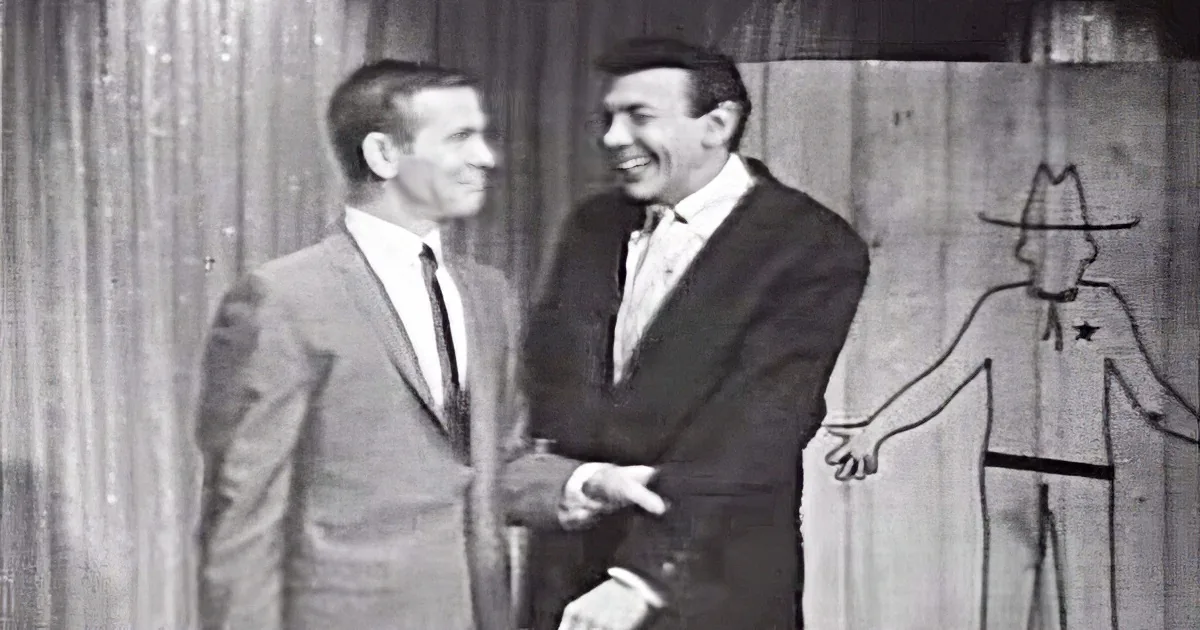 Two men in suits shake hands, smiling, against a rustic wooden backdrop with a stick figure.