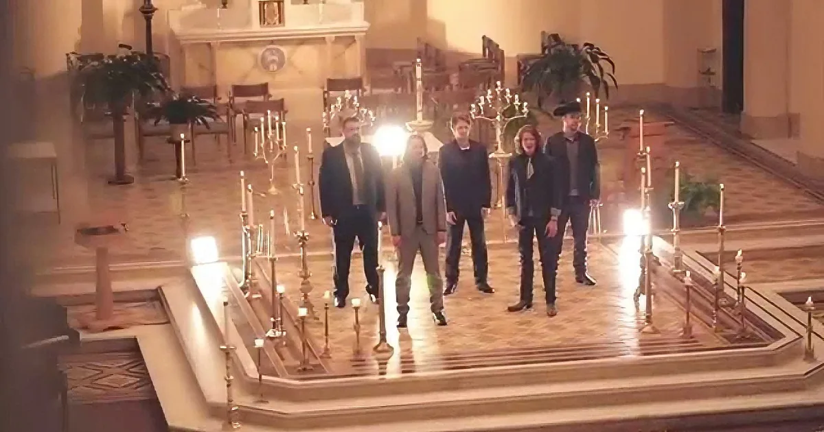 A group of six people in formal attire stands on a church altar surrounded by lit candles.
