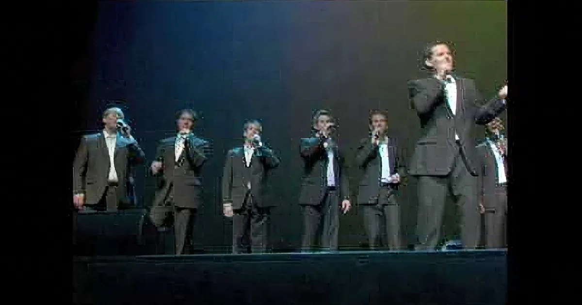 A group of men in suits sing on stage, illuminated by stage lights.
