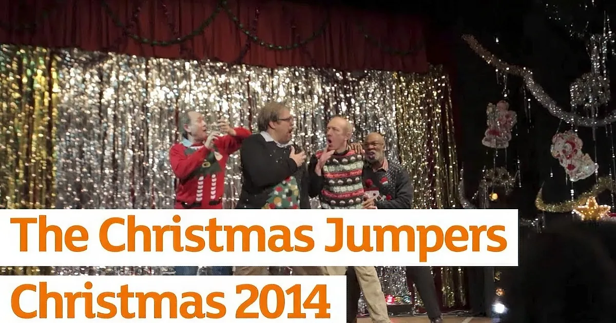 Four men in festive sweaters perform on stage with a Christmas backdrop and decorations.