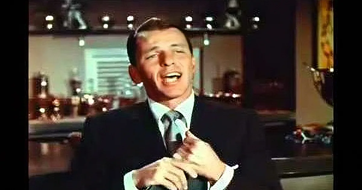 A man in a suit and tie is singing passionately, with a microphone in front of him, in a studio setting.