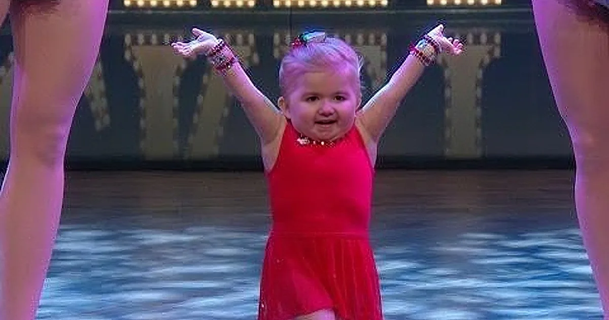 A young child in a red dress is dancing with arms raised, wearing bracelets and a necklace.