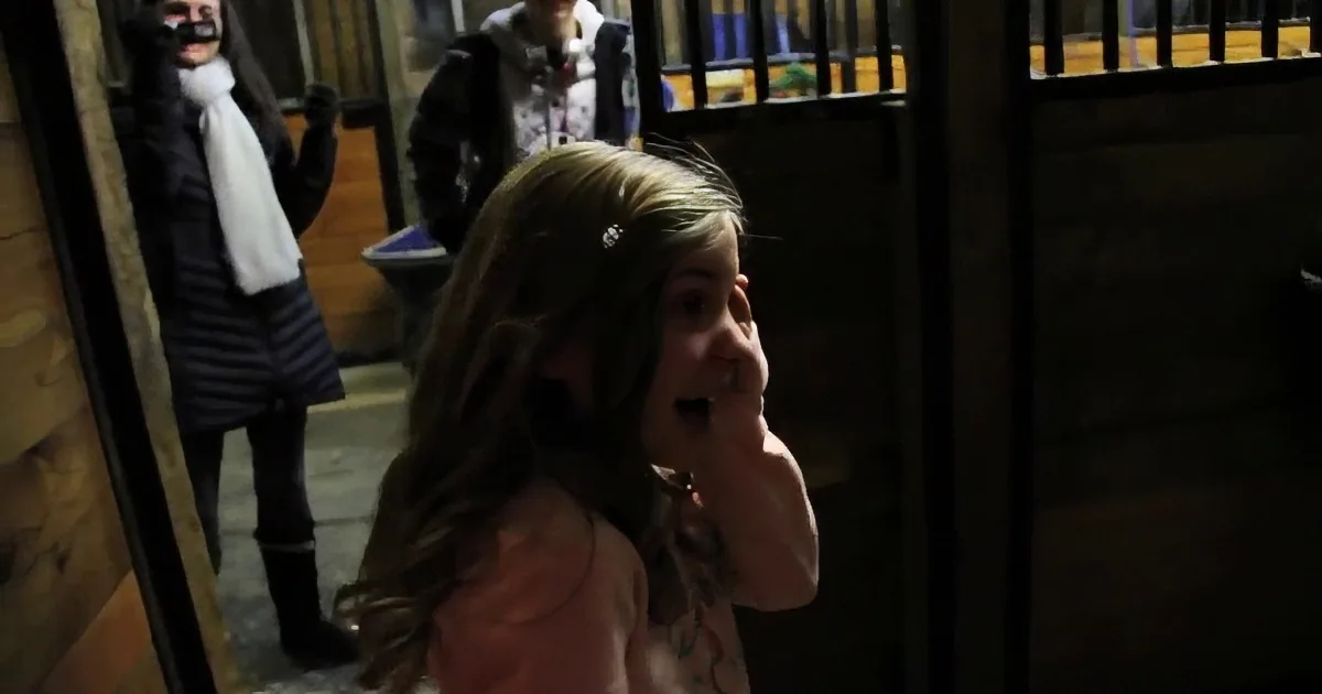 A young girl with long hair, wearing a pink top, stands in a dimly lit area, looking to the side.