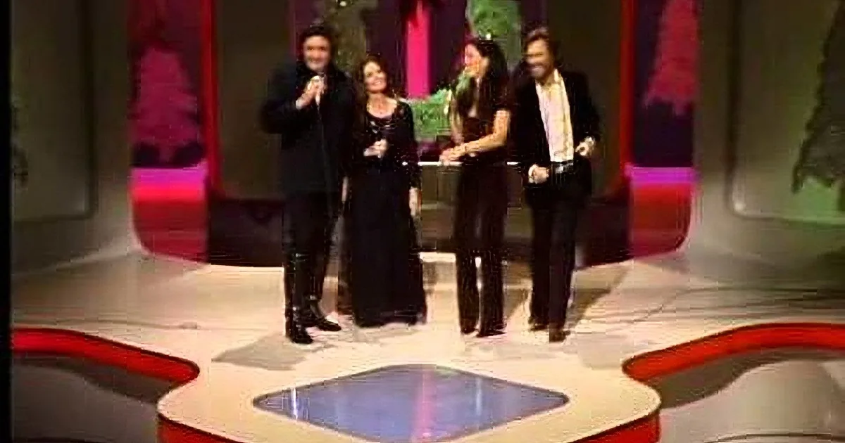Four people stand on a stage with a festive backdrop, one holding a microphone.