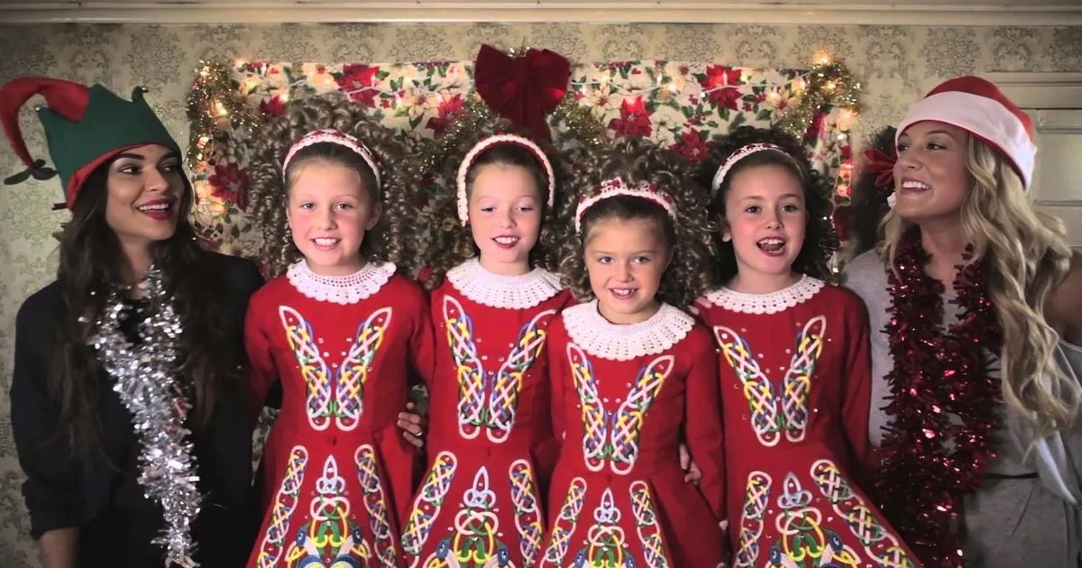 Five young girls in red dresses with intricate designs, smiling, flanked by two women in festive attire, against a...