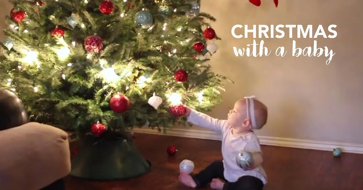 A baby plays with ornaments near a decorated Christmas tree while an adult assists.