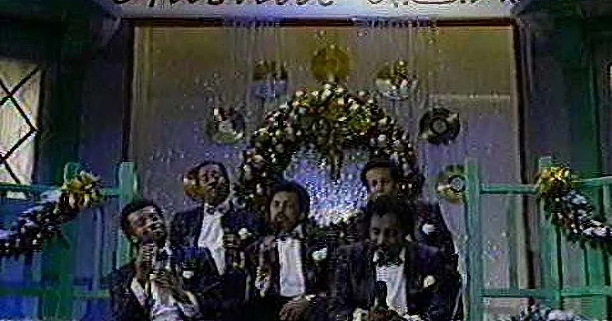 A group of men in formal attire sing on stage with a floral arrangement and decorations behind them.