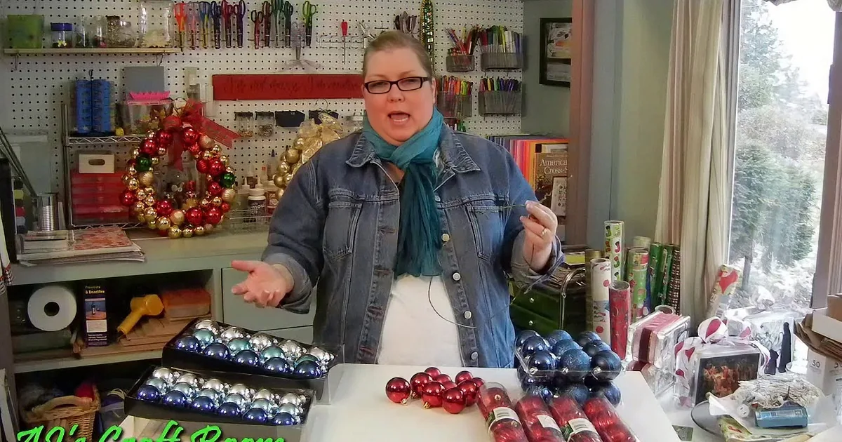 A woman in a craft room arranges Christmas ornaments on a table, surrounded by crafting supplies and decorations.