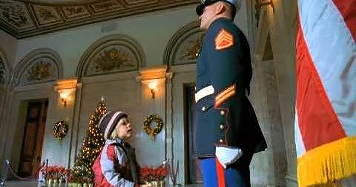 A young child in a red jacket stands in front of a Christmas tree, looking up at a uniformed officer in a grand room.