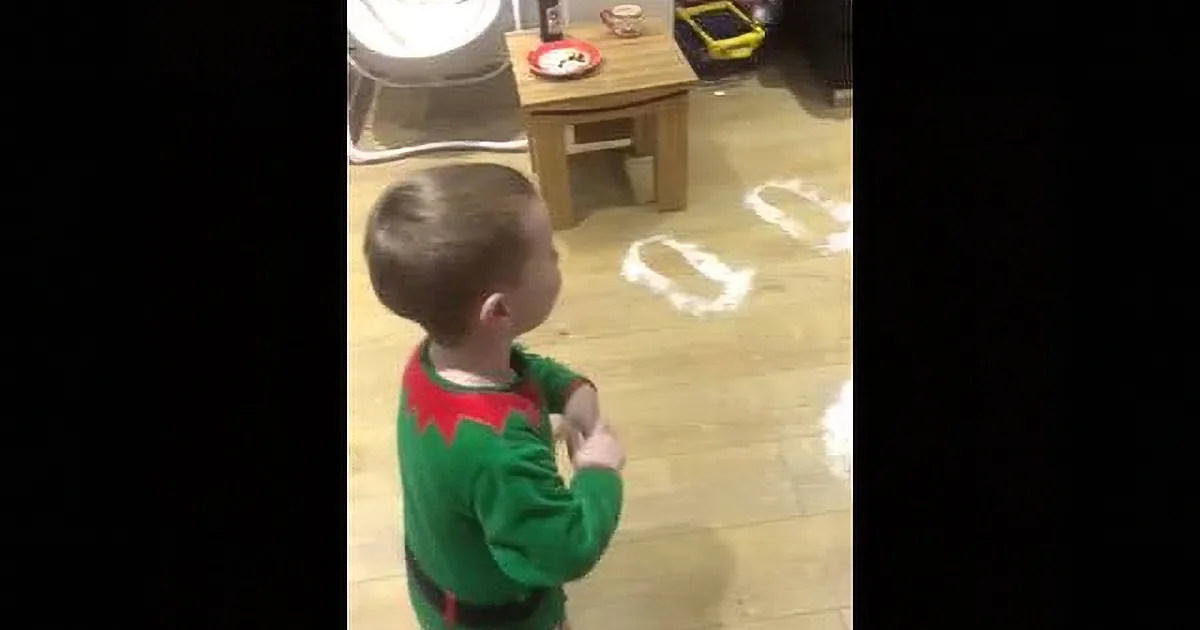 A child in a green Christmas outfit stands facing a table with a red plate, while white footprints are drawn on the floor.