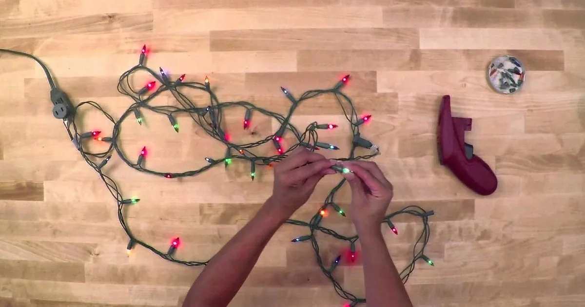 Hands untangle Christmas lights on a wooden floor near a vacuum cleaner and outlet.