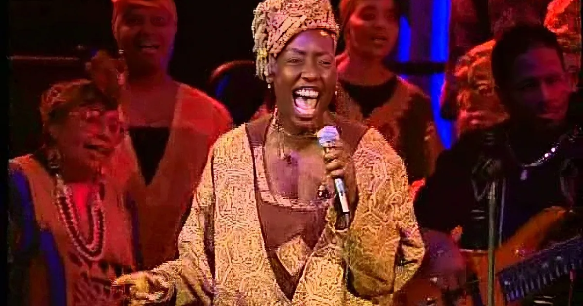 A woman in a patterned outfit sings into a microphone, surrounded by a group of people in colorful attire.