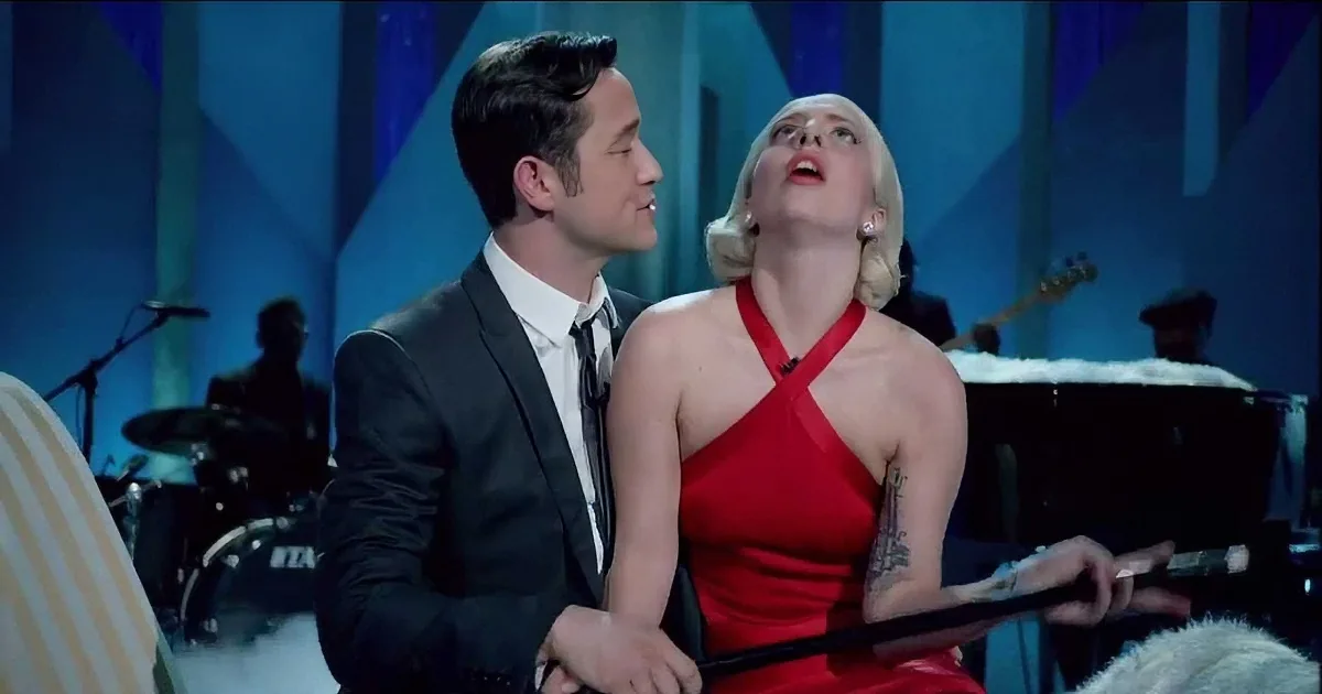 A man in a suit and a woman in a red dress are seated together, with the woman holding a microphone and looking up.