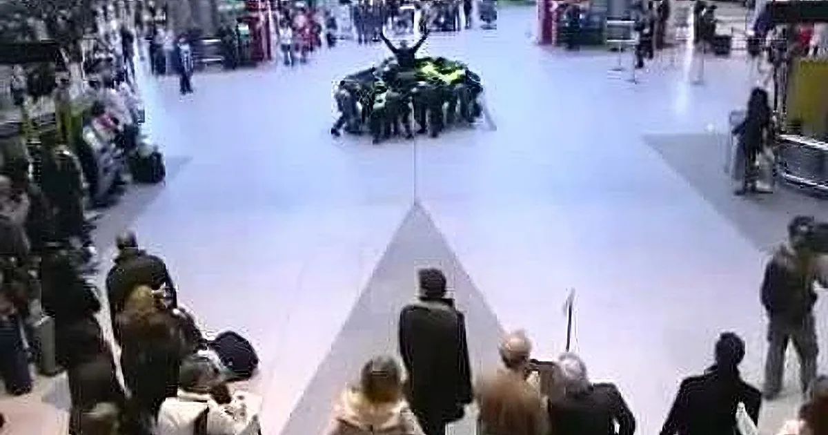 A group of people in a circle, possibly performing or dancing, surrounded by onlookers in a spacious indoor area.