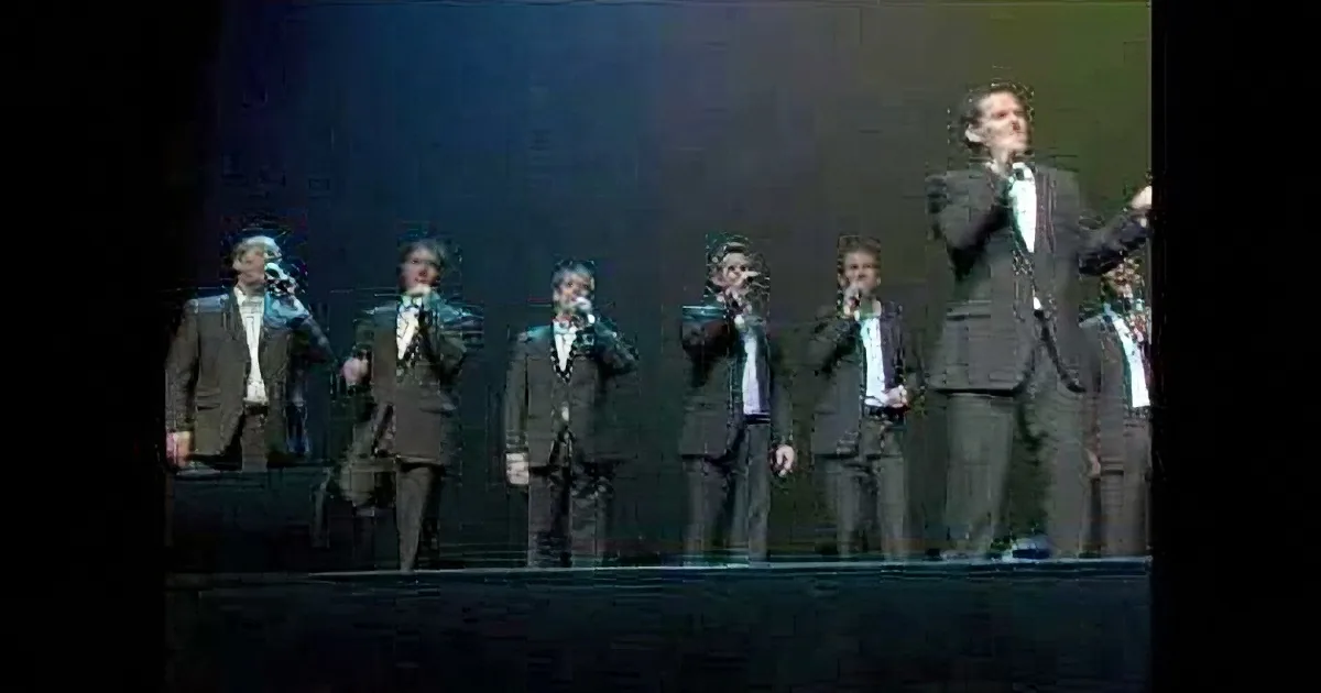 A group of men in suits sing on stage under dim lighting.